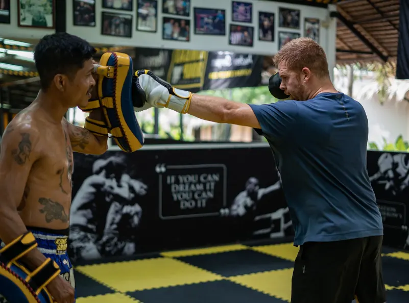 A beginner Muay Thai student practicing punches with a trainer holding pads at Kombat Group in Thailand.