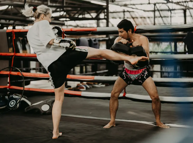 A Muay Thai practitioner delivering a powerful kick during a pad training session with a trainer at Kombat Group Thailand. The image captures the intensity and focus of the training environment in the gym's boxing ring.