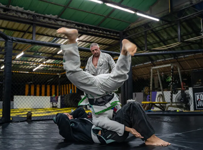 Two men practice Brazilian Jiu-Jitsu with one executing a sweep on the other, under the watchful eye of their instructor at Kombat Group in Pattaya, Thailand. The image captures the intensity and skill development during the training session, highlighting the dedicated instruction and dynamic environment of the BJJ training camp.