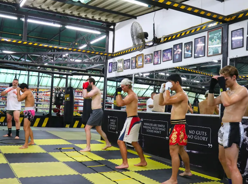 A group of students practicing Muay Thai techniques in a training session at Kombat Group Thailand, surrounded by a professional gym setup and motivational quotes on the walls.