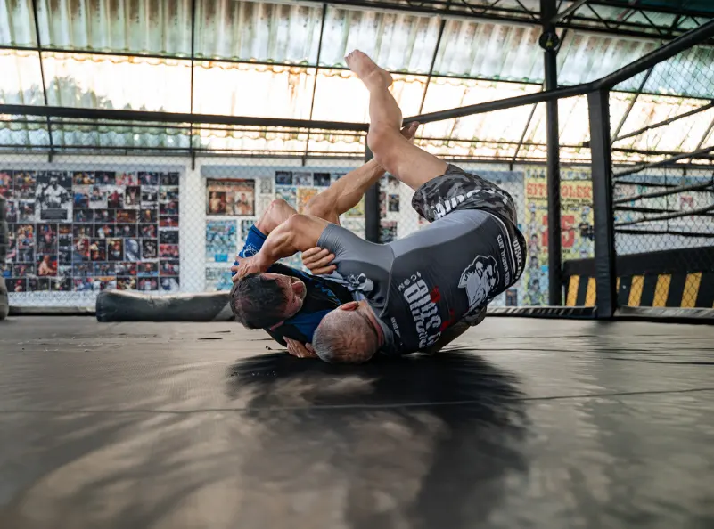 Two men engage in an intense Brazilian Jiu-Jitsu no GI rolling session at Kombat Group in Thailand. The scene captures the dynamic action and focus of their training, with the gym's vibrant background adding to the energetic atmosphere. This image showcases the dedication and skill development fostered at the BJJ training camp.