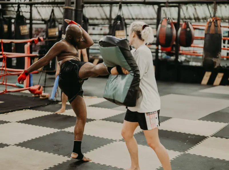 A male Muay Thai student delivering a powerful roundhouse kick to a female student holding a pad. The training takes place at Kombat Group’s state-of-the-art facility, where participants build strength, technique, and confidence through Muay Thai.