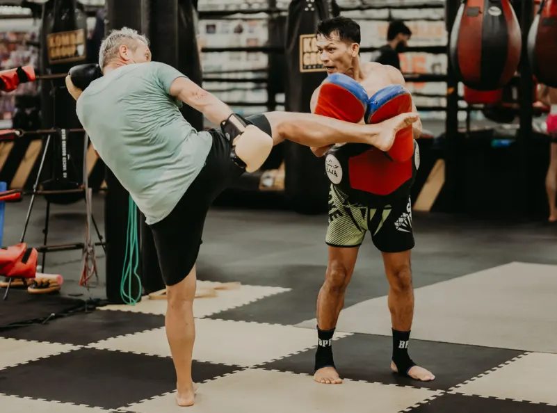 An older student executes a powerful kick on a coach’s pads during a training session at Kombat Group Thailand, focusing on technique and power.